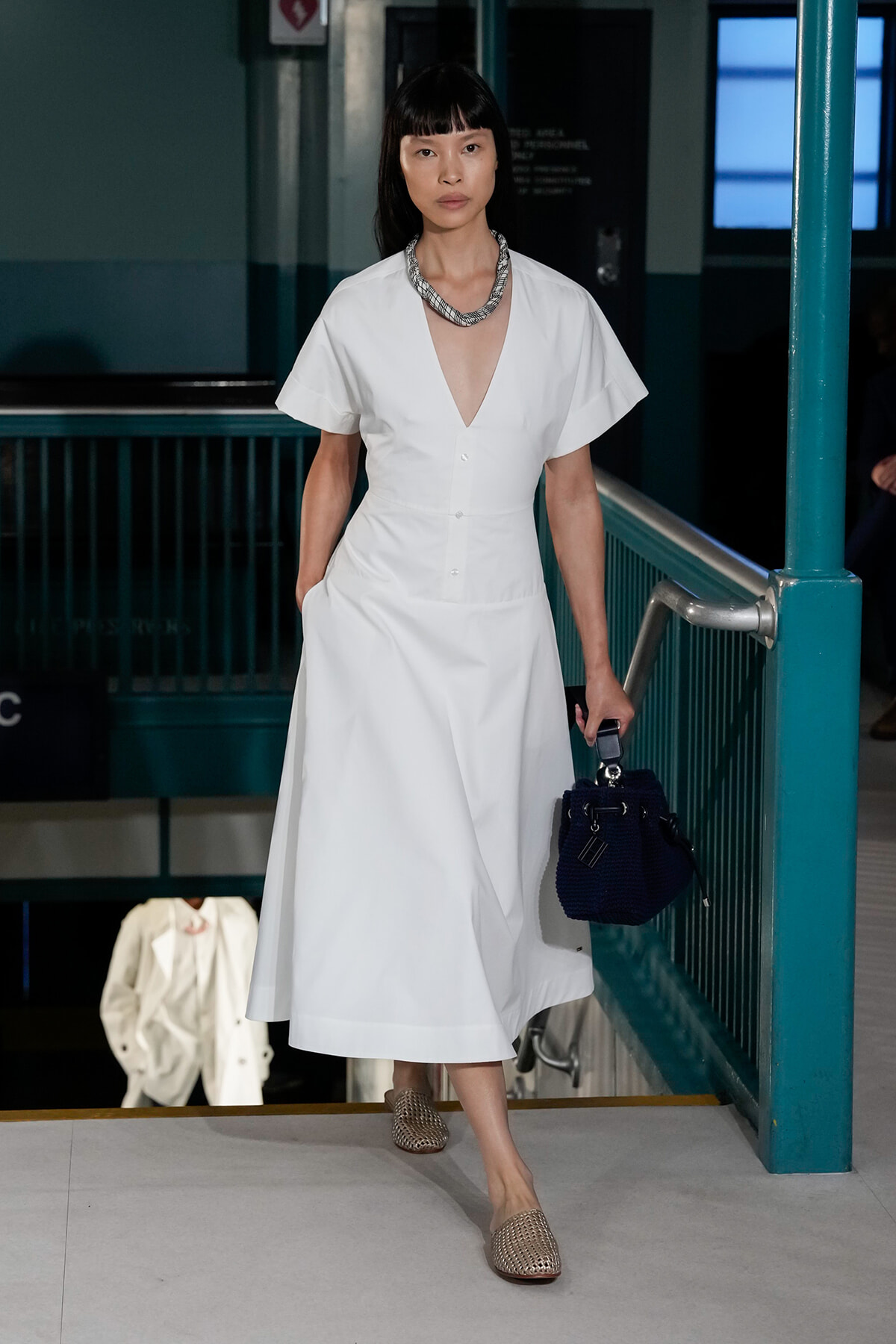 Model in a white V-neck midi dress with short sleeves walks down a runway stair, carrying a navy handbag and wearing a chunky necklace.