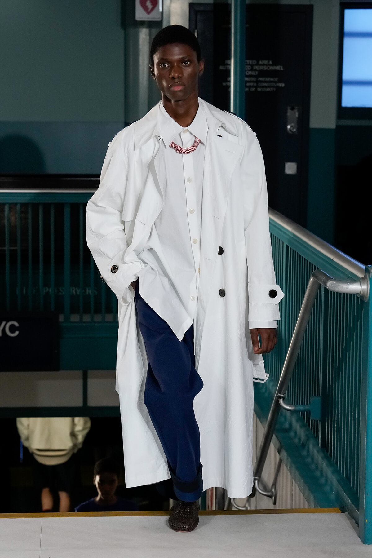 Male model in an oversized white trench coat over a white shirt and blue pants, walking down a runway staircase in a fashion show setting.