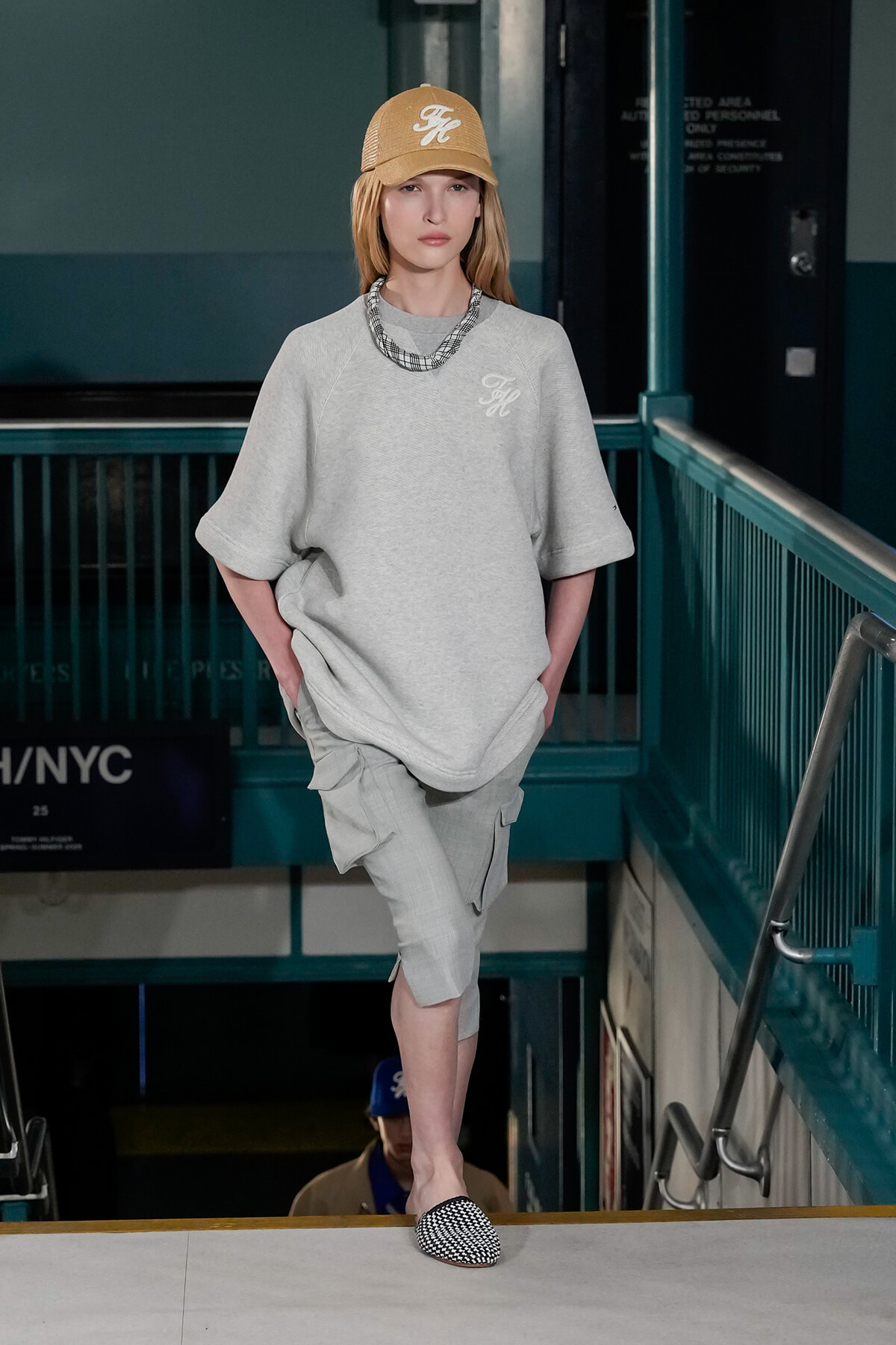 Model on a runway wearing a gray oversized top, light cargo shorts, tan cap, and black-and-white checkered loafers in a subway-style venue