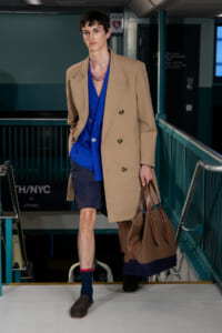 Male model in a beige double-breasted trench over a bright blue shirt and navy shorts, carrying a large brown tote bag while walking down a stairwell.