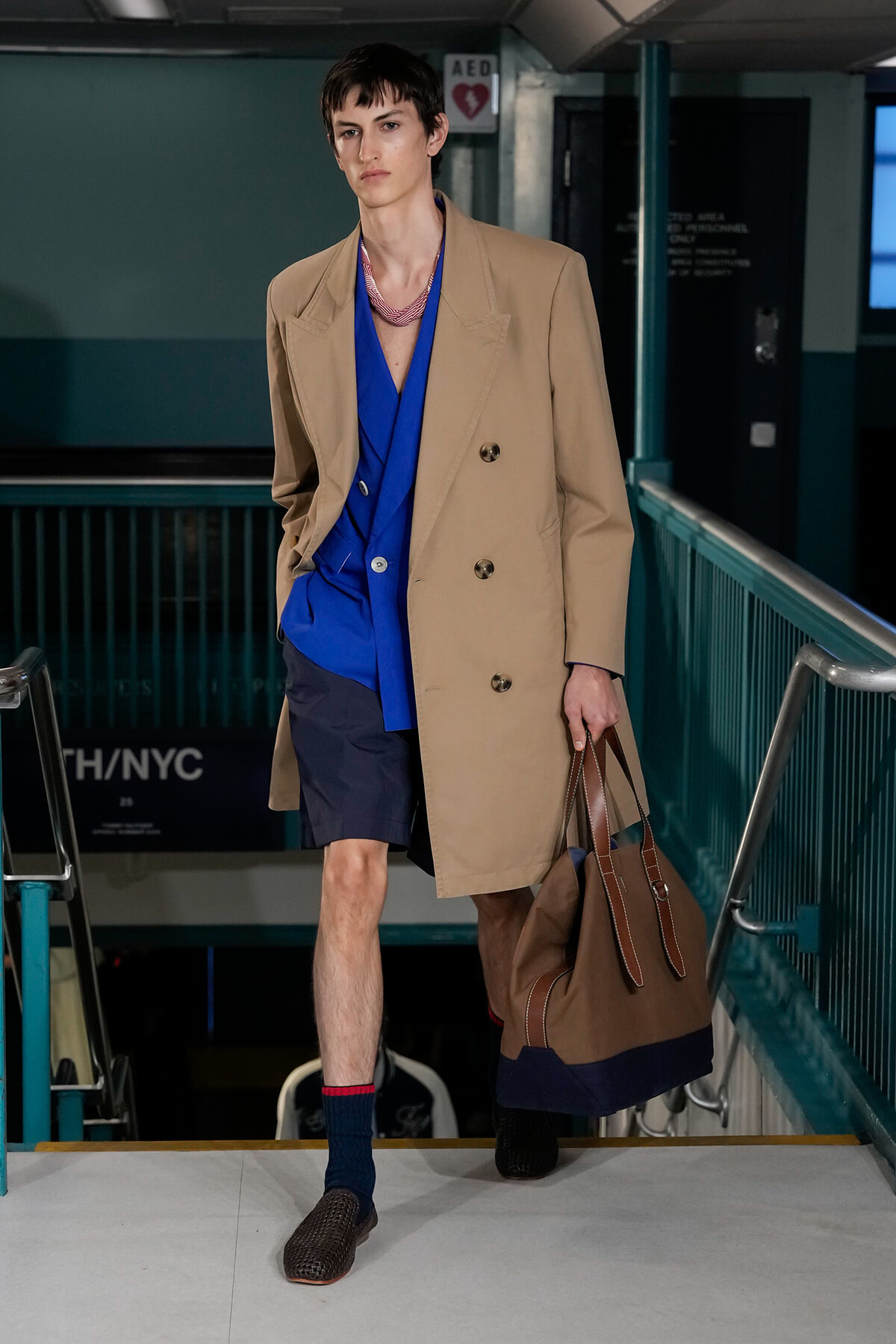 Male model in a beige double-breasted trench over a bright blue shirt and navy shorts, carrying a large brown tote bag while walking down a stairwell.