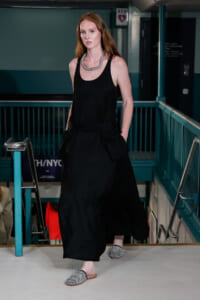 Woman in a sleeveless black dress walking down a stairwell, wearing a chunky silver necklace and black-and-white checkered flats.
