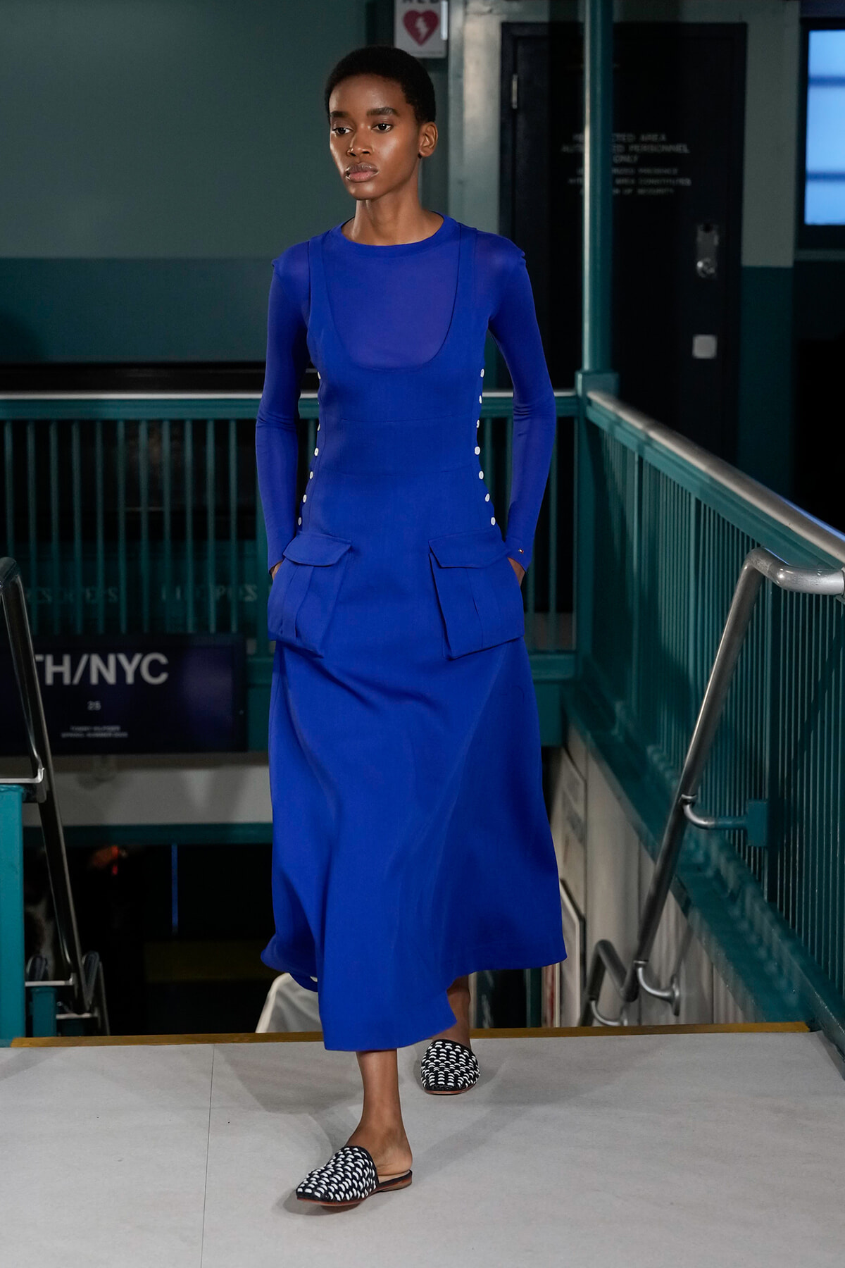 Female model in a cobalt blue long-sleeve dress with oversized front pockets and white side buttons, walking a runway in a teal-toned indoor venue with patterned slip-ons.