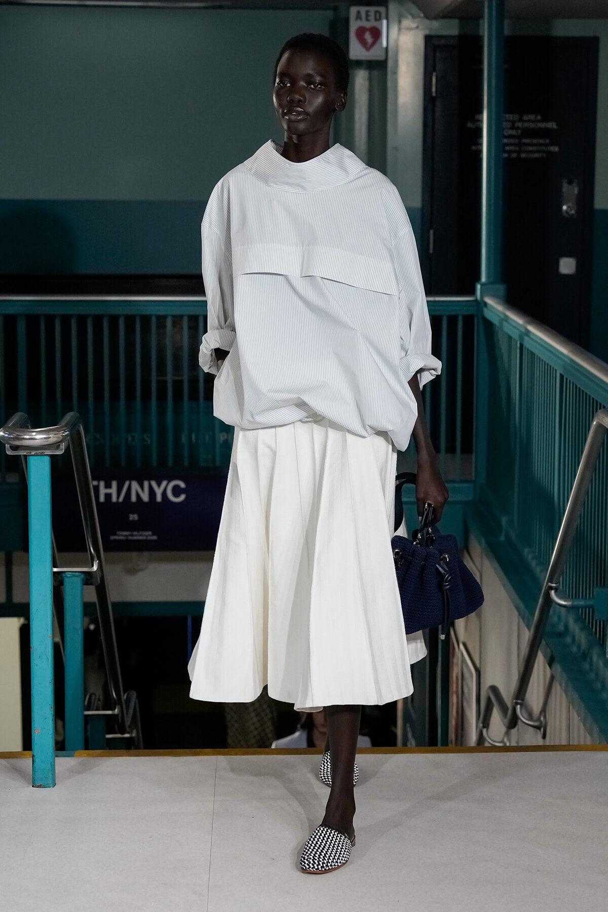 Black model walks a runway in an oversized white top and white midi skirt, carrying a navy handbag; checkered flats complete the look in a subway-station setting.