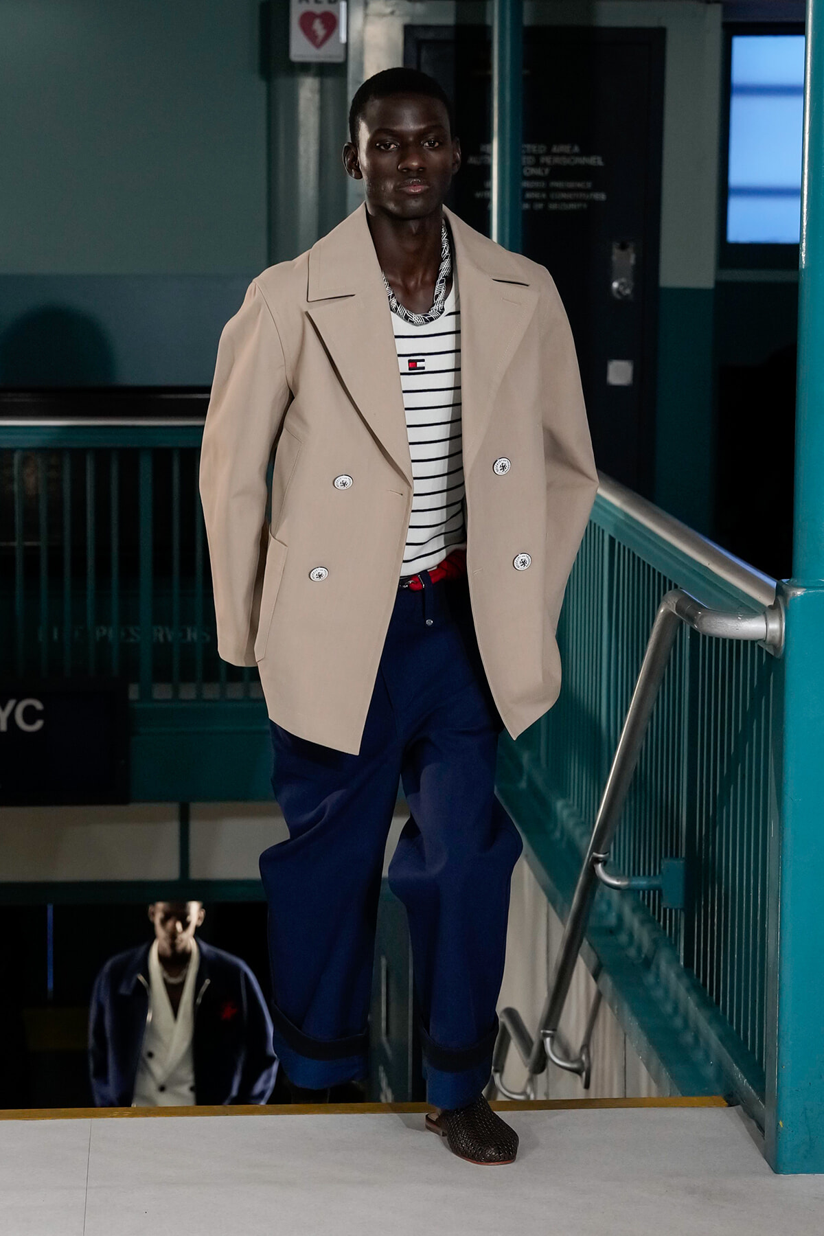 Man wearing a beige double-breasted coat over a striped shirt descends a subway stairwell in blue pants and brown shoes.