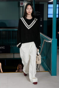 Model walking down a runway stair in a teal subway setting, wearing an oversized black knit sweater with white chevron neckline, white wide-leg pants, red shoes, and carrying a beige woven bag.