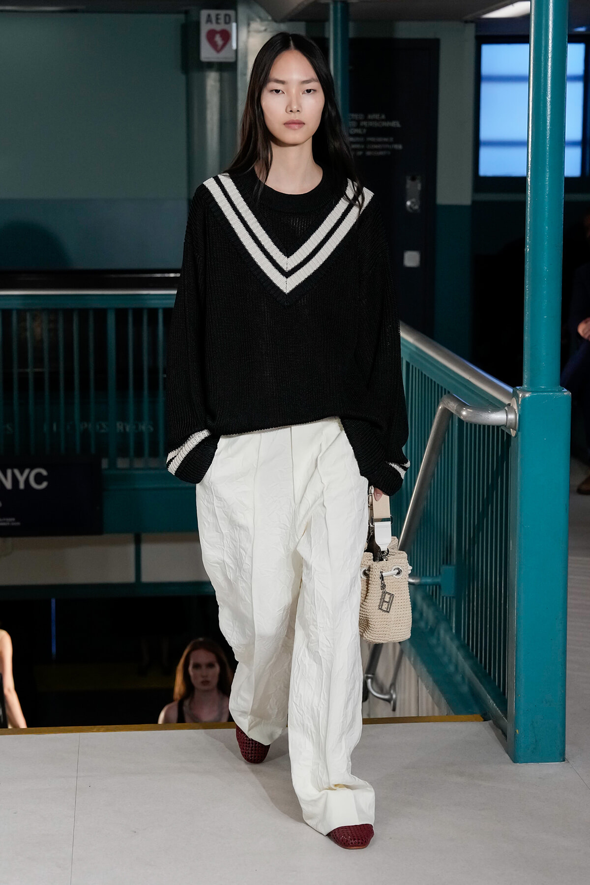 Model walking down a runway stair in a teal subway setting, wearing an oversized black knit sweater with white chevron neckline, white wide-leg pants, red shoes, and carrying a beige woven bag.