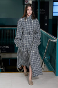 Model wearing an oversized navy plaid trench coat with a belt, walking down stairs in a subway-like setting.