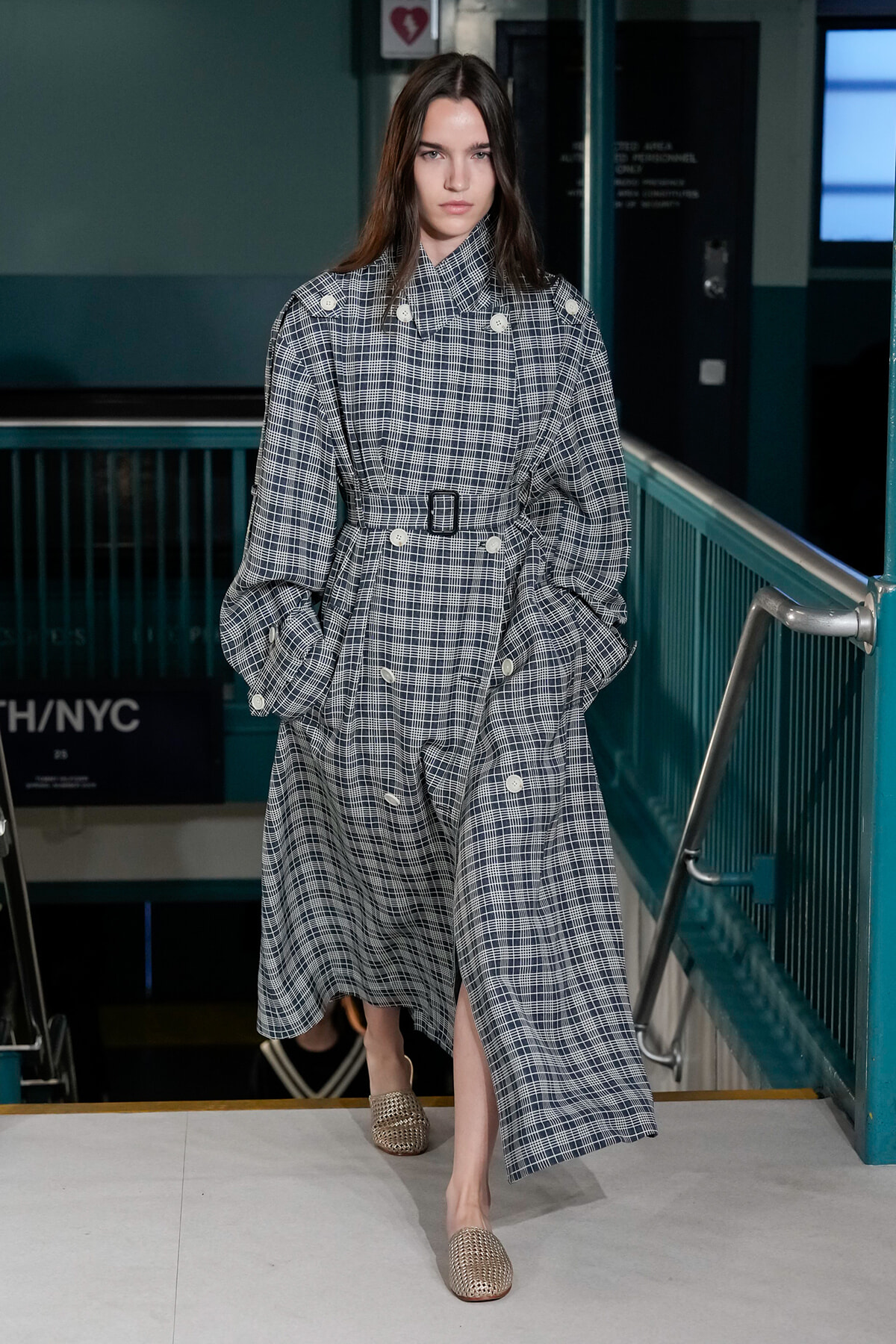 Model wearing an oversized navy plaid trench coat with a belt, walking down stairs in a subway-like setting.