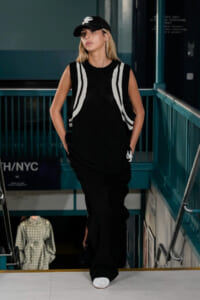 Female model on a runway wearing a black sleeveless knit top with white stripe details, black pants, and a black cap with a white logo.