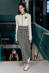 Model walking in a cream cardigan with black buttons and navy script, paired with black-and-white plaid wide-leg pants and white oxford shoes on a runway.