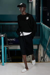 Person wearing a black knit sweater with a white logo, black shorts, and white perforated slides in a subway stairwell. Cap and hands in pockets, looking aside.