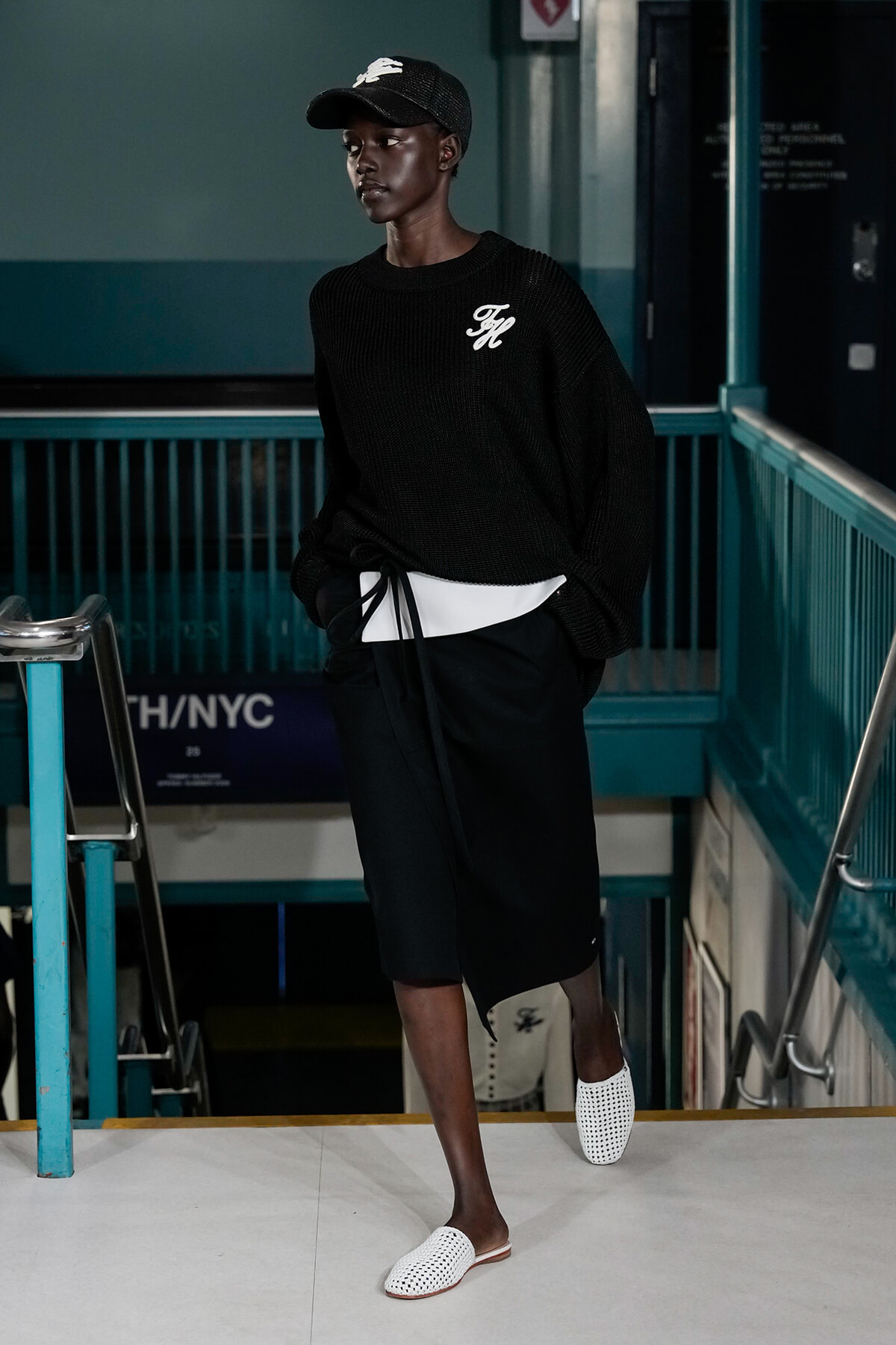 Person wearing a black knit sweater with a white logo, black shorts, and white perforated slides in a subway stairwell. Cap and hands in pockets, looking aside.