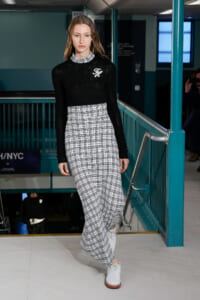 Model walking runway in a black knit cropped sweater over a white plaid long dress with white shoes in a teal subway-station setting
