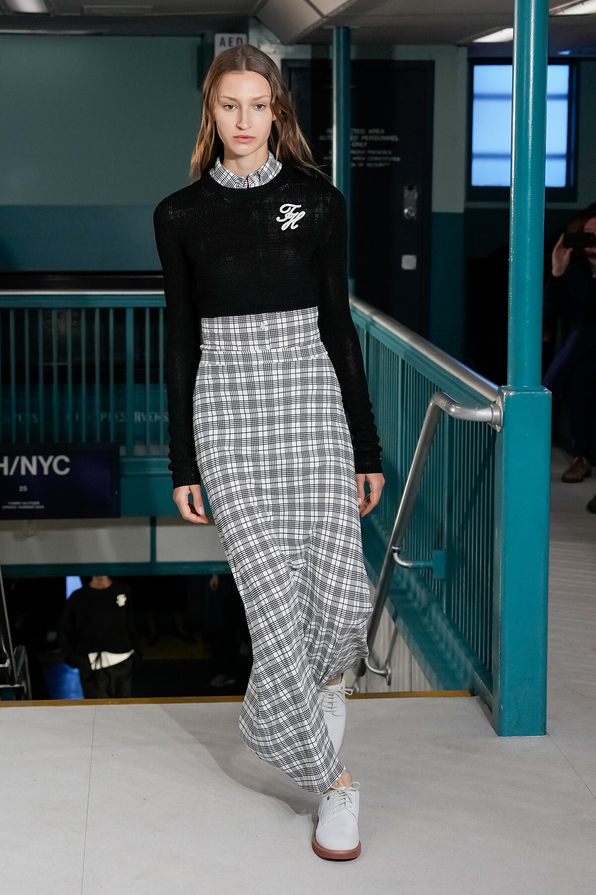 Model walking runway in a black knit cropped sweater over a white plaid long dress with white shoes in a teal subway-station setting