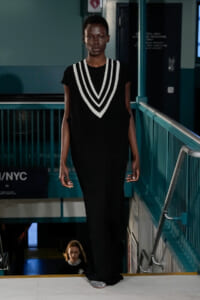 Model walking runway in a black knit dress with bold white V-neck stripes, teal railings in the background, and a footstep-ready pose.