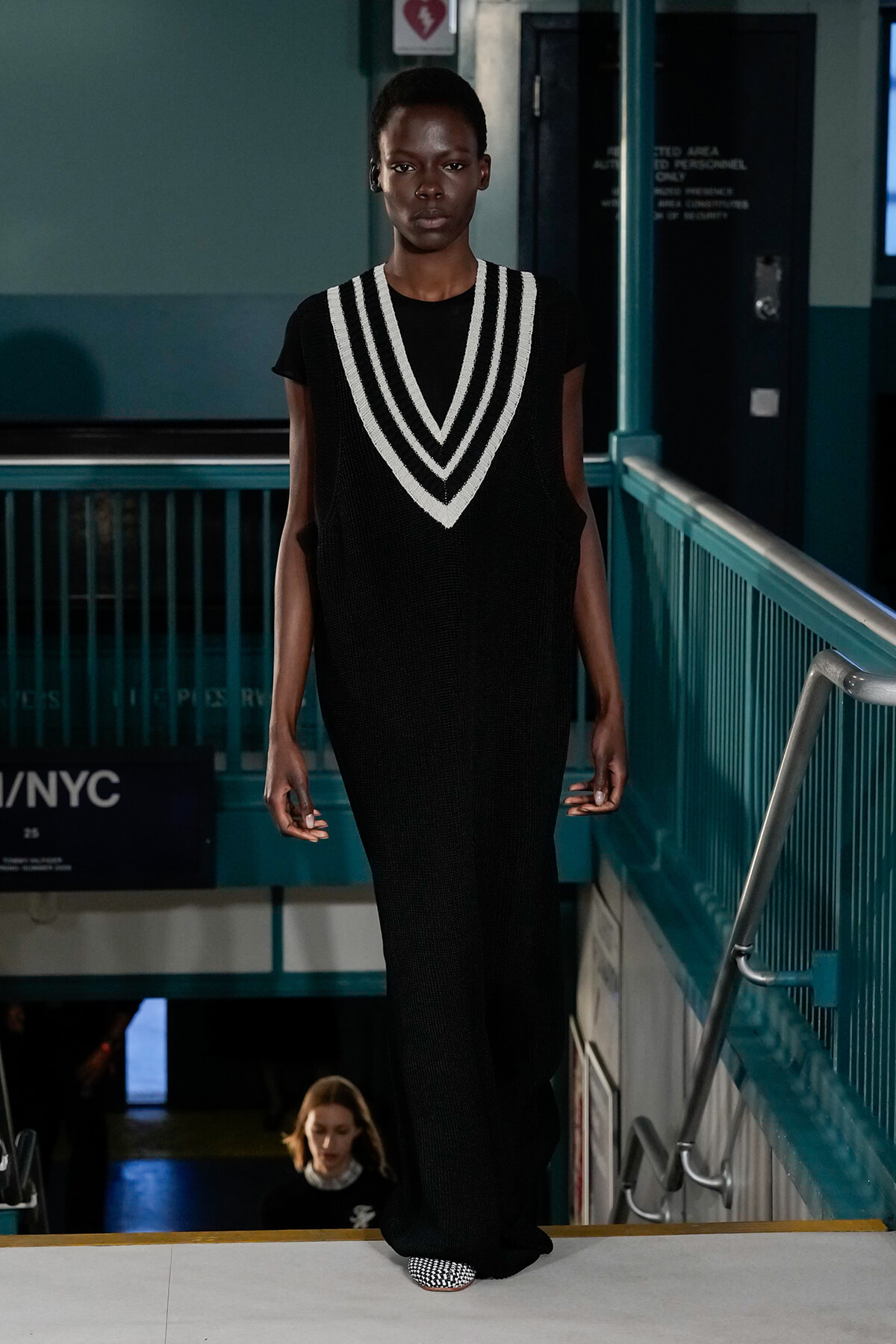 Model walking runway in a black knit dress with bold white V-neck stripes, teal railings in the background, and a footstep-ready pose.