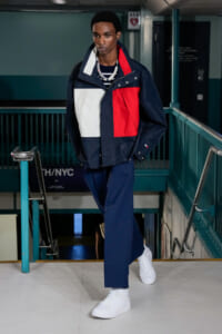 Male model walking down a backstage stairwell in a navy, white and red color-block jacket, navy trousers, and white sneakers.