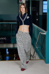 Female model walks a runway wearing a navy cropped cardigan with white trim and plaid wide-leg pants, red textured shoes, in an urban backstage setting.