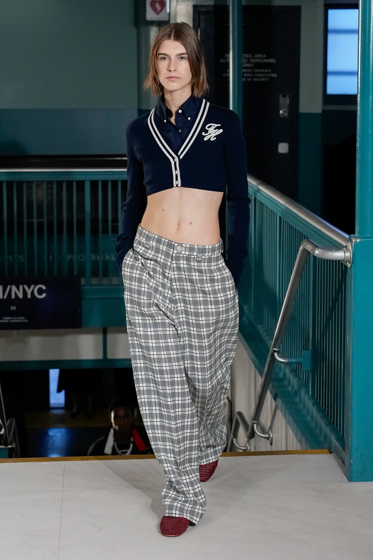 Female model walks a runway wearing a navy cropped cardigan with white trim and plaid wide-leg pants, red textured shoes, in an urban backstage setting.