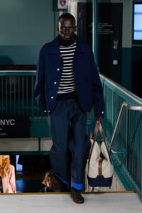 Male model in a navy jacket and striped sweater walks down a subway stairwell, carrying a beige and navy tote bag.