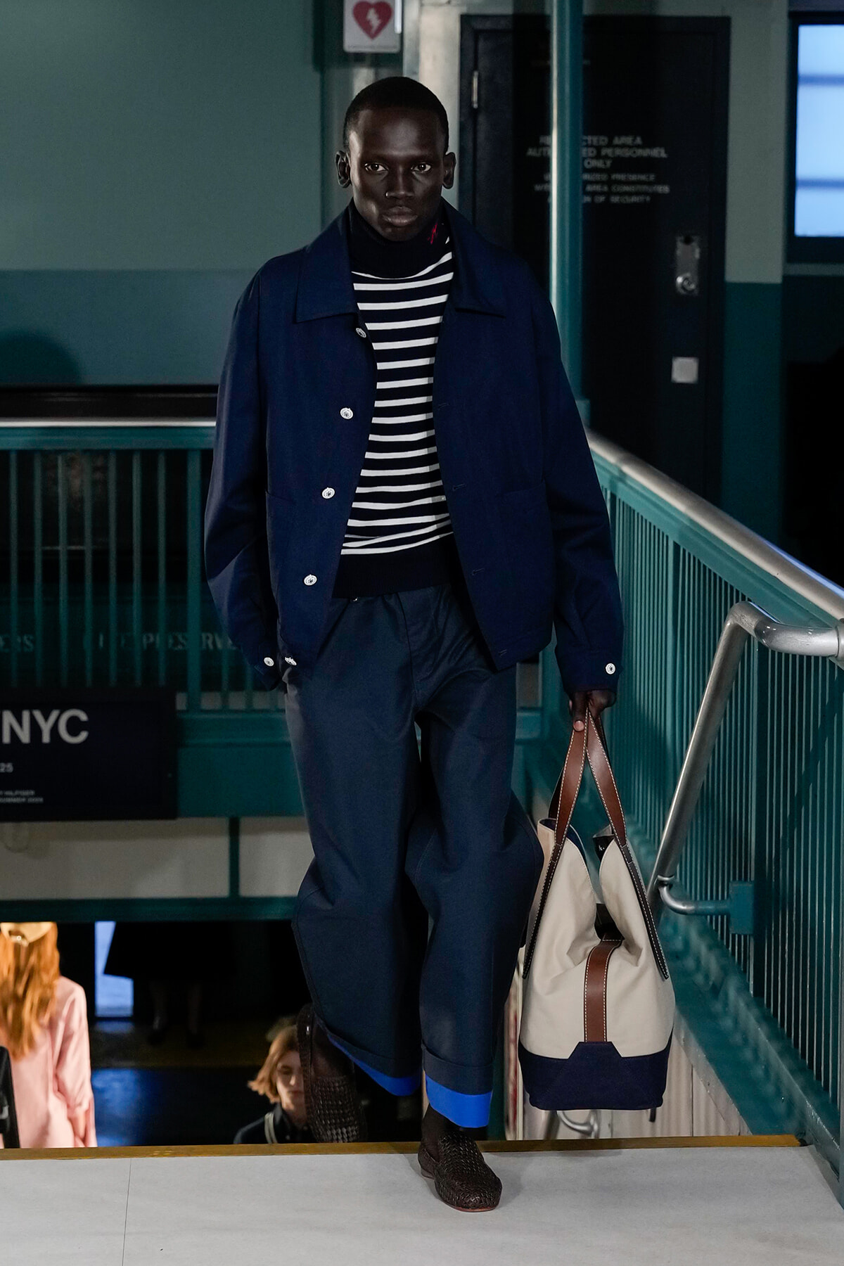 Male model in a navy jacket and striped sweater walks down a subway stairwell, carrying a beige and navy tote bag.