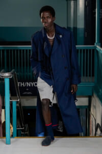 Male fashion model walks down a stair-step runway in a long navy trench coat, wearing a silver layered necklace.