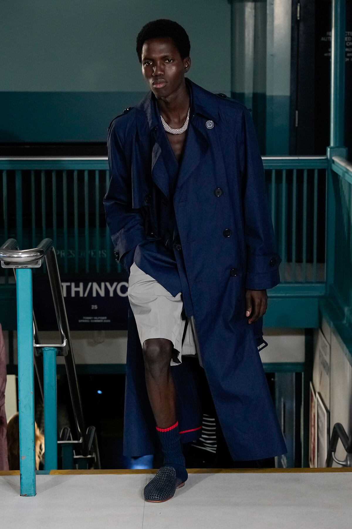 Male fashion model walks down a stair-step runway in a long navy trench coat, wearing a silver layered necklace.