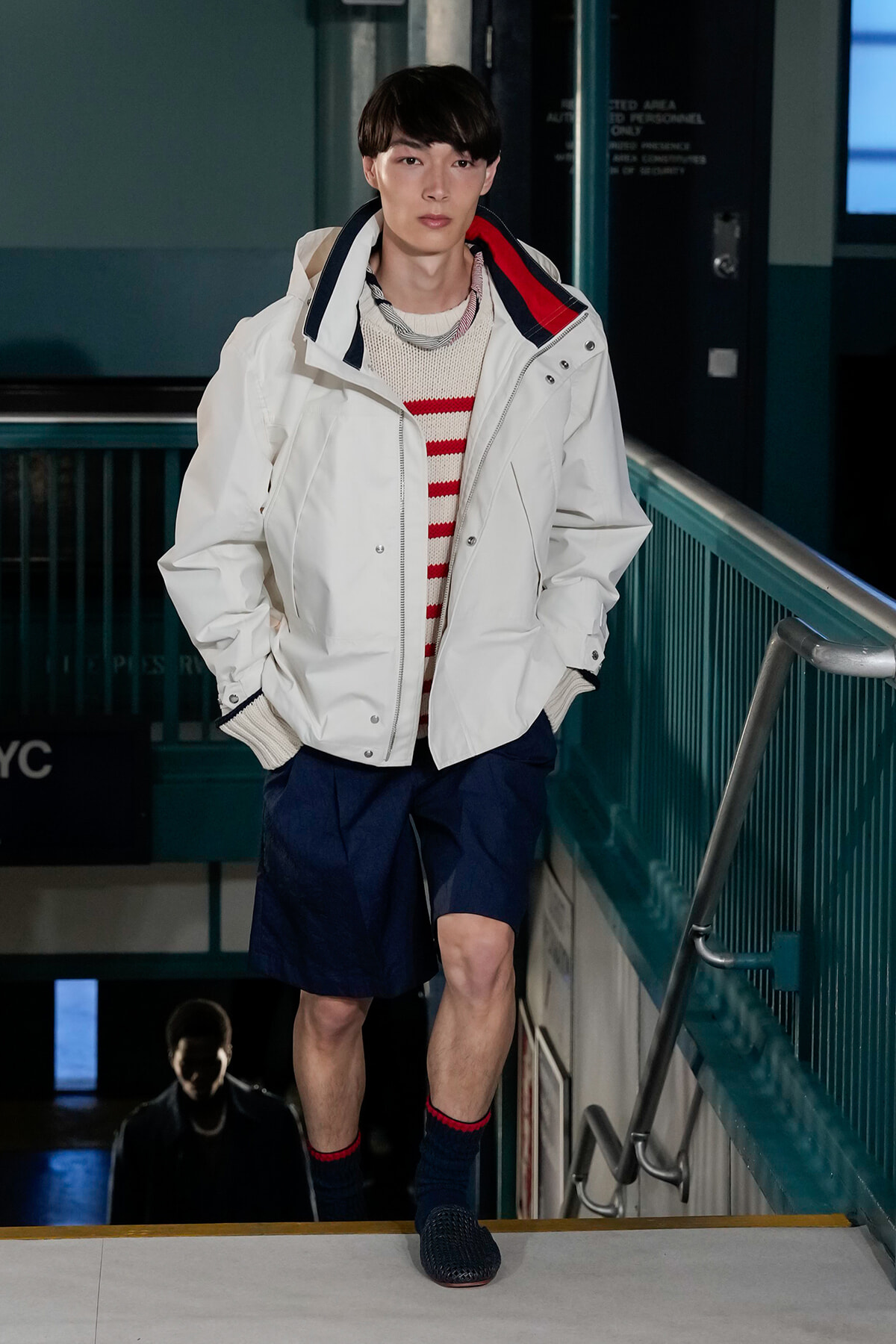Male model wearing a white jacket over a red-and-white striped sweater and navy shorts descends a stairwell in a subway station, hands in pockets.