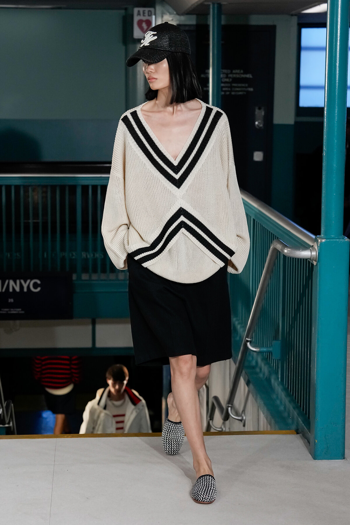 Model wearing an oversized cream sweater with bold black V stripes, black shorts, and a black cap, walking on a subway-style runway.