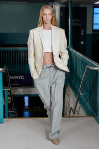 Model walking a fashion runway in a cream blazer, white cropped top, and light gray tailored pants inside an industrial venue.