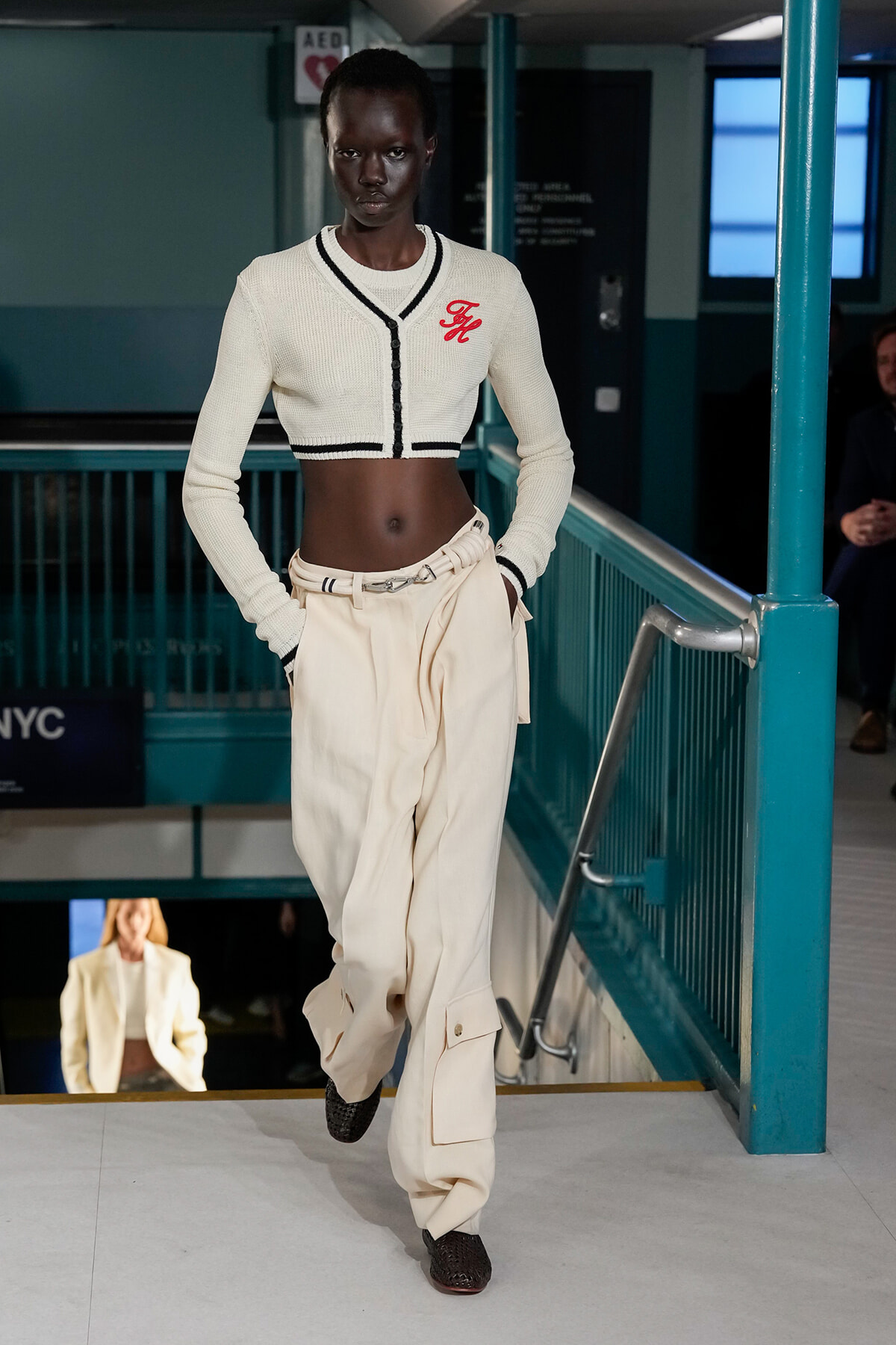 Model walking down a runway in a cream cropped cardigan with black trim and high-waisted cream cargo pants, black shoes, in a teal subway-station set.