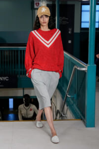 Model walking a fashion show in a red knit sweater with a white chevron V, light gray knee-length shorts, white woven loafers, and a beige cap.