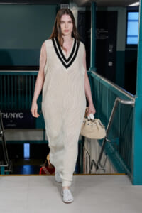 Model walking on a runway in an oversized cream knit sleeveless vest with black and white V-neck trim, carrying a beige woven handbag.
