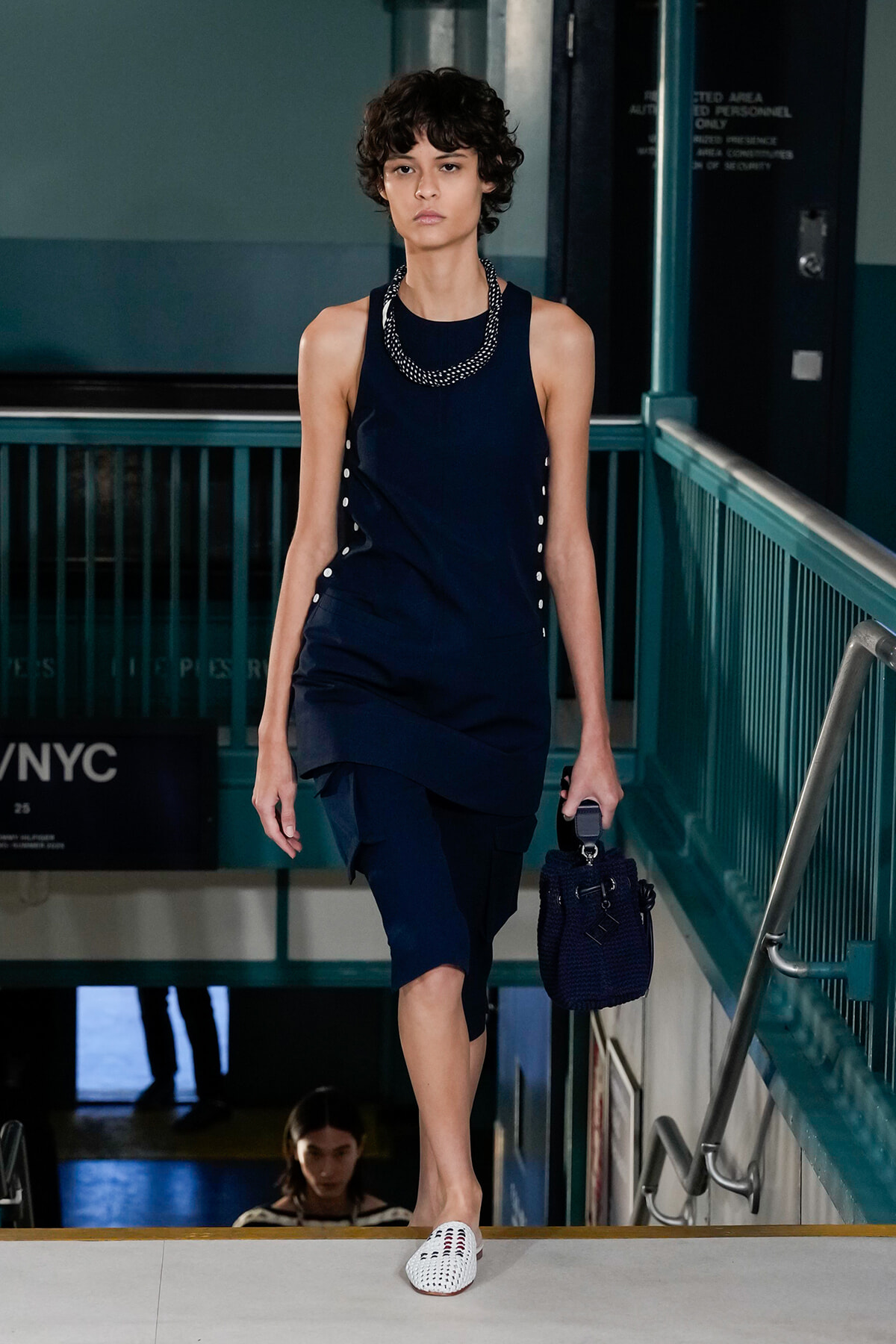 Model walks a runway in a navy sleeveless outfit with side buttons, layered hem, chunky beaded necklace, and navy bag with white woven flats.