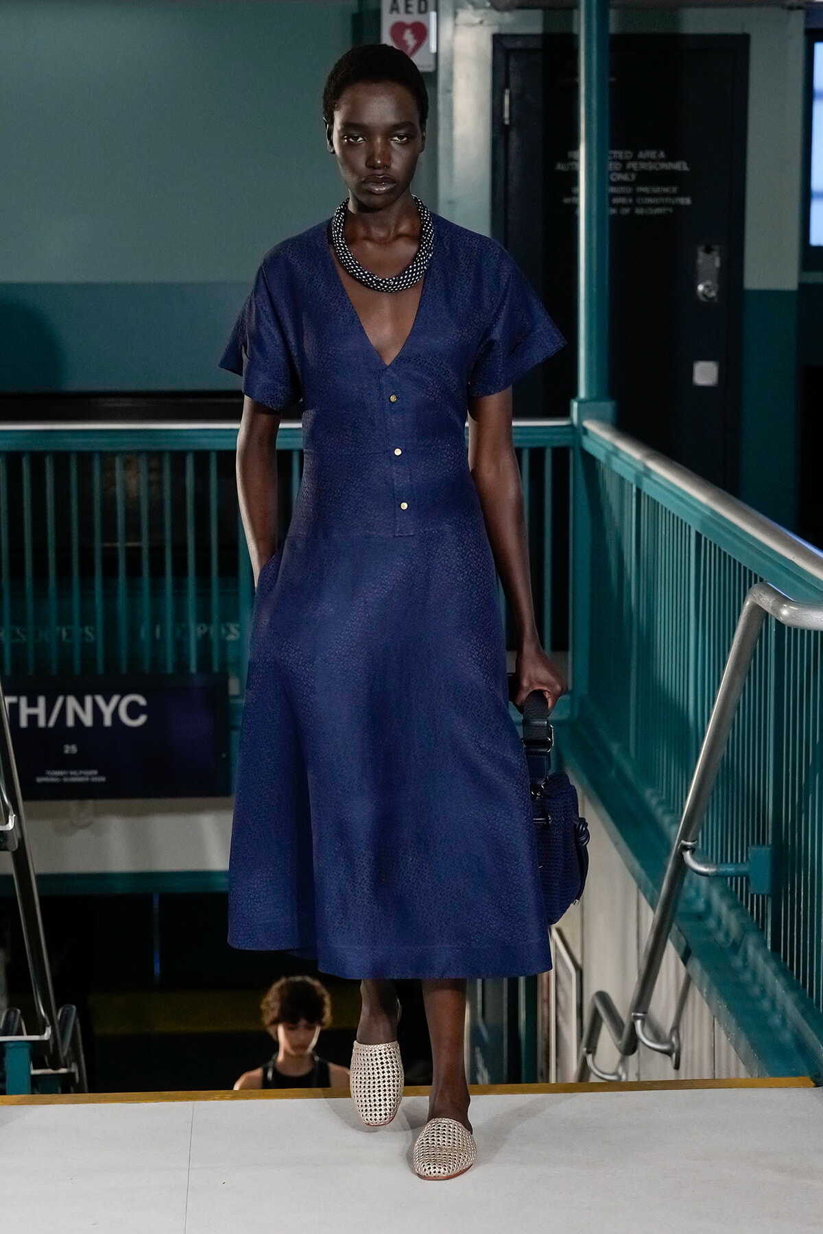 Model walking a runway in a textured cobalt blue midi dress with a deep V-neck and button placket, accessorized with a chunky black-and-white necklace and white woven mules.