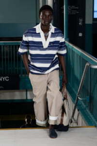 Man walking down a teal staircase wearing a navy and white striped polo, beige cuffed pants, and carrying a bag.