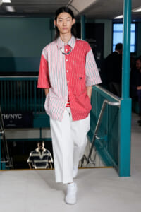 Model walking on a runway in a red-and-white split-pattern shirt with white wide-leg pants and white sneakers.