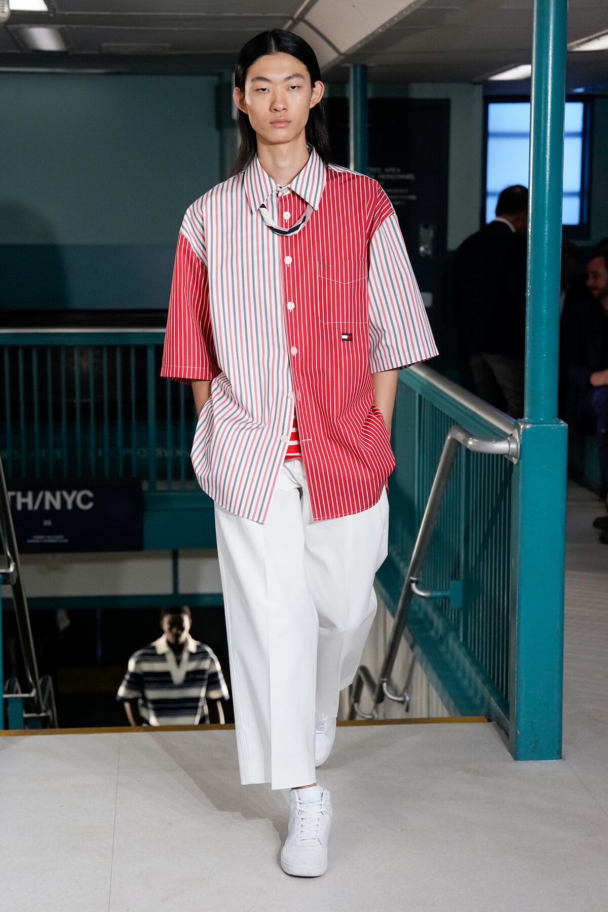 Model walking on a runway in a red-and-white split-pattern shirt with white wide-leg pants and white sneakers.