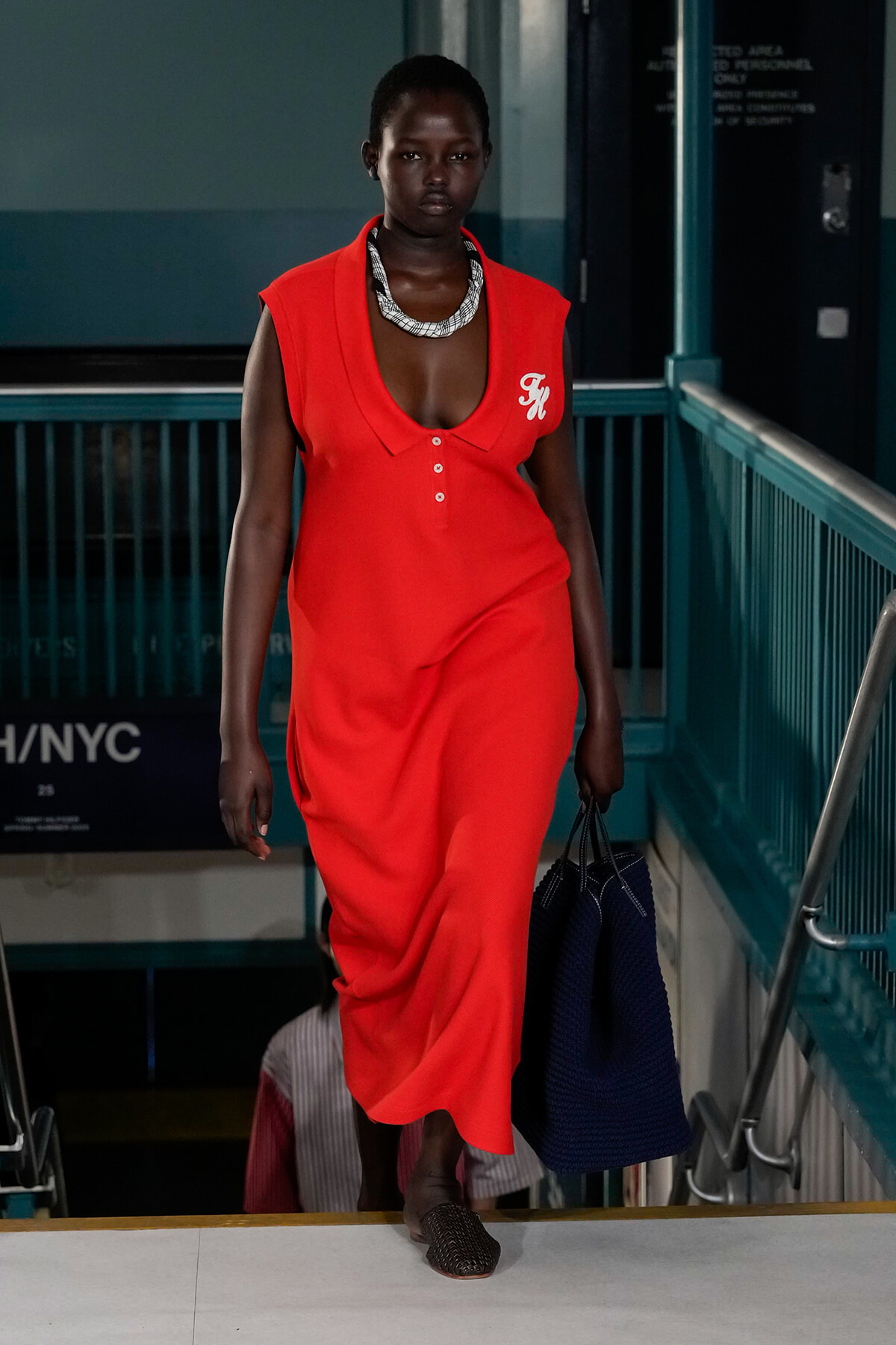 Woman in a bright red sleeveless dress with a white logo, descending stairs in a transit area while carrying a dark blue bag.