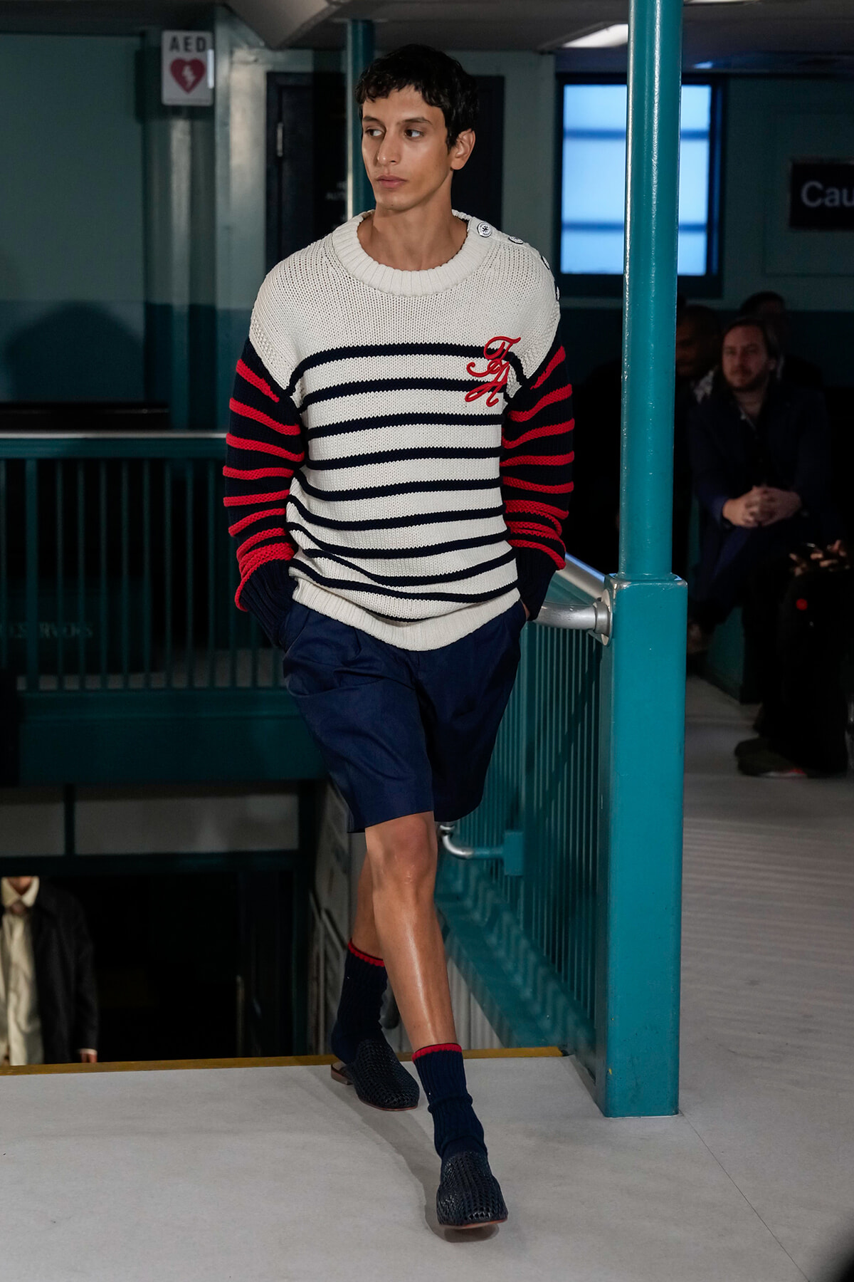 Male model wearing a white knit striped sweater with navy and red stripes, navy shorts, and navy socks walking down a runway staircase in a teal-colored venue.