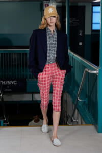 Model walking down a runway in an urban/subway-style setting, wearing a navy blazer, checkered shirt, red plaid cargo shorts, white woven shoes, and a tan cap.