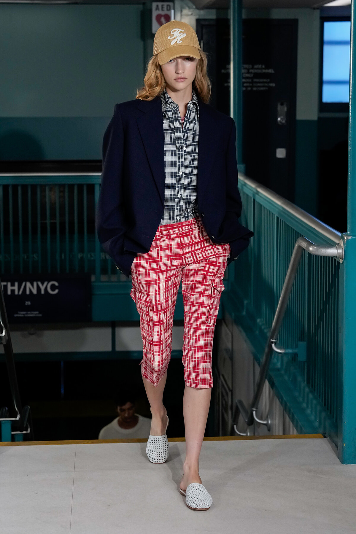 Model walking down a runway in an urban/subway-style setting, wearing a navy blazer, checkered shirt, red plaid cargo shorts, white woven shoes, and a tan cap.