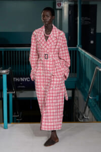 Model wearing a pink plaid trench coat with belt, standing on a runway indoors.