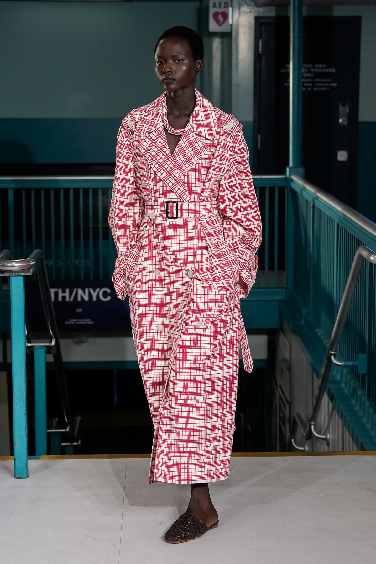 Model wearing a pink plaid trench coat with belt, standing on a runway indoors.