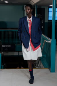 Male model on a runway wearing a navy double-breasted blazer over a red striped shirt, white skirt-like shorts, and navy socks with red trim.