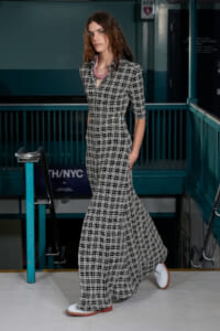 Woman in a black-and-white plaid maxi dress walking on a subway platform, wearing white shoes with brown soles.
