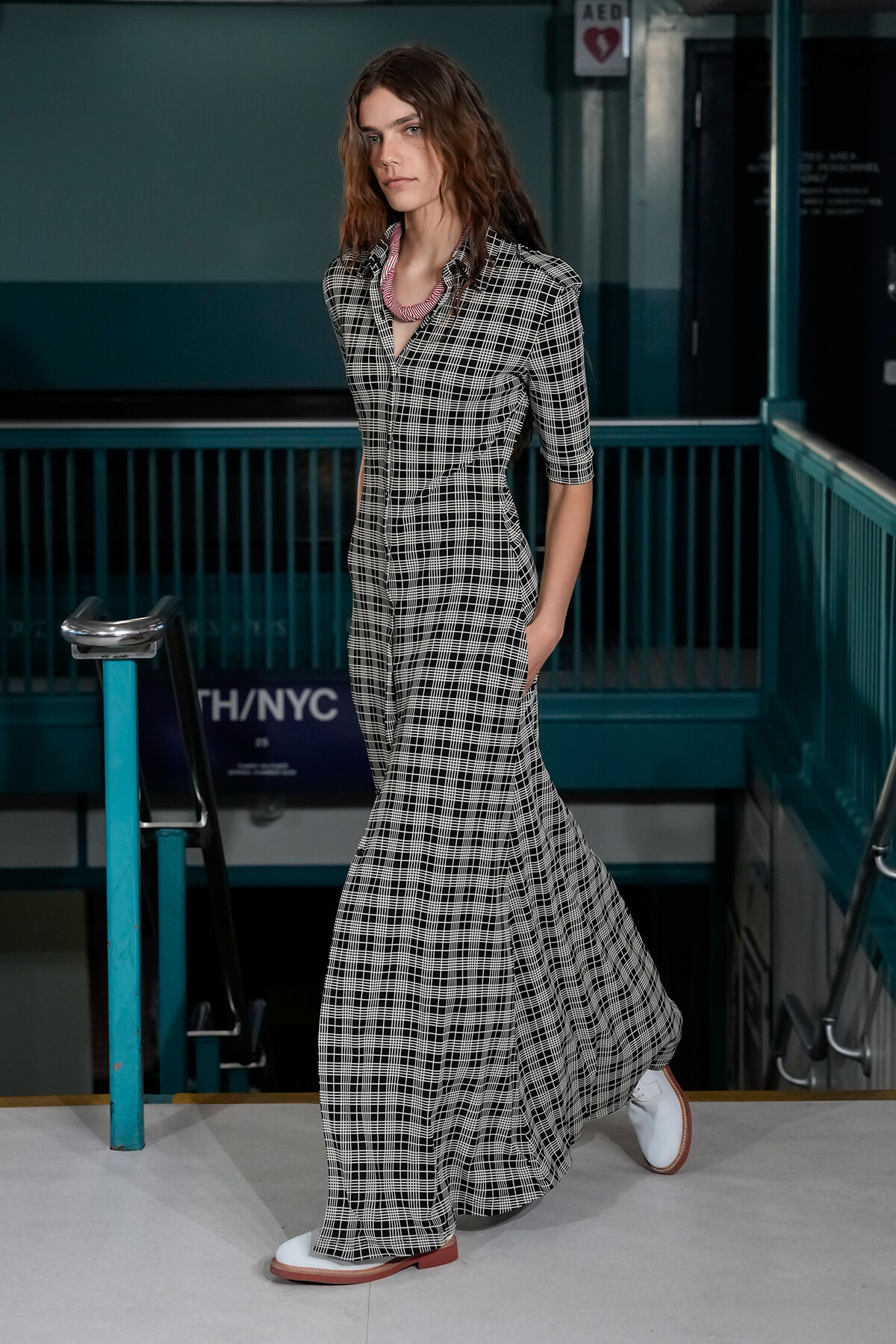 Woman in a black-and-white plaid maxi dress walking on a subway platform, wearing white shoes with brown soles.