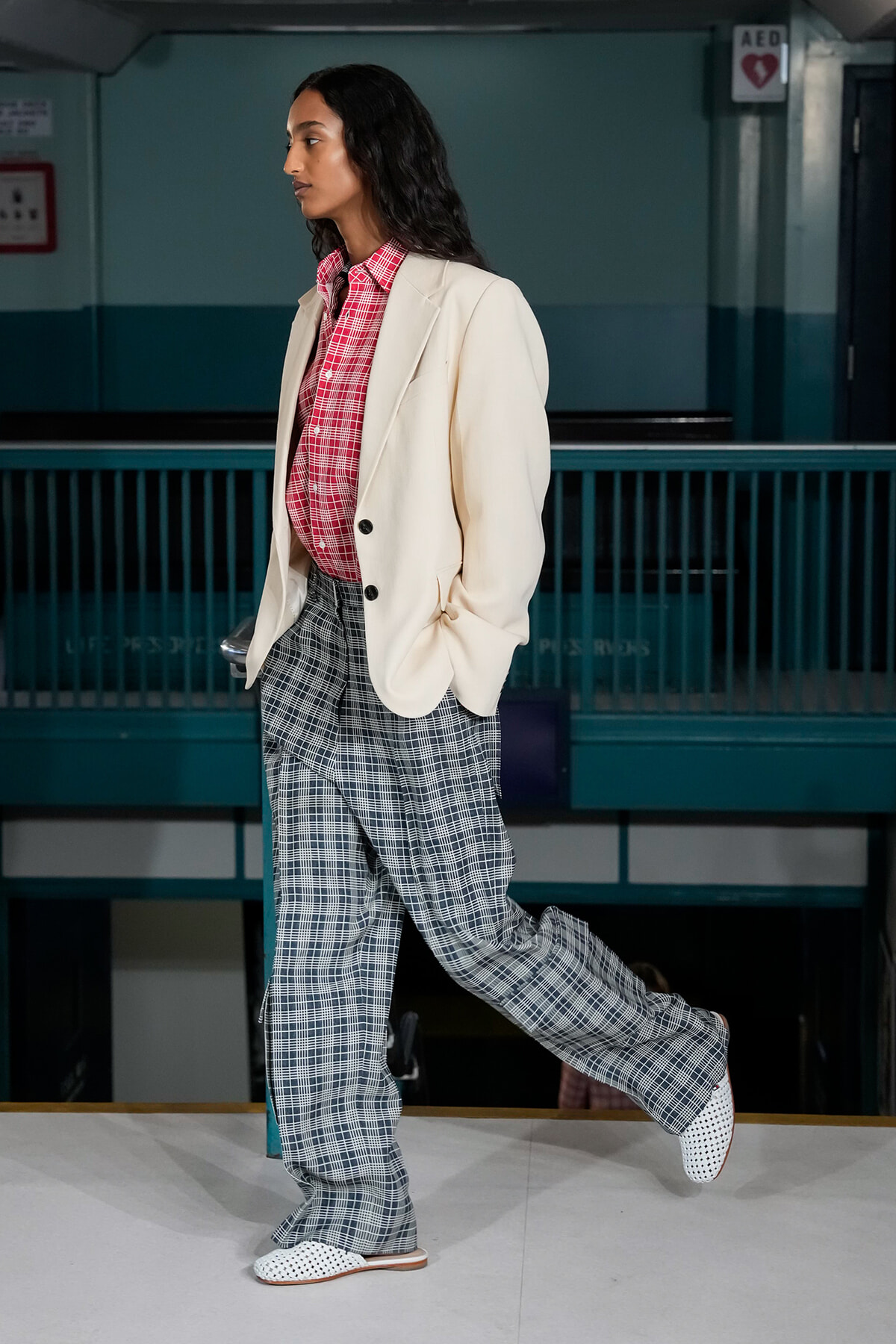 Model walking in a subway station wearing a cream blazer over a red plaid shirt and gray plaid wide-leg pants with white dotted shoes.