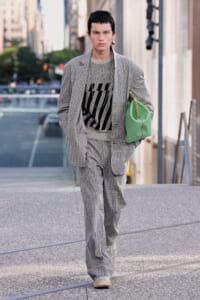 Fashion model in a gray checkered suit and patterned sweater walking down a city street, carrying a green handbag under his arm.